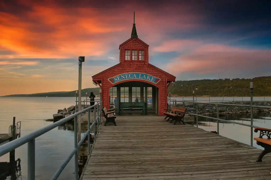 Wooden pier leading to a red pavilion on Seneca Lake in Watkins Glen, New York under a vibrant sunset sky.