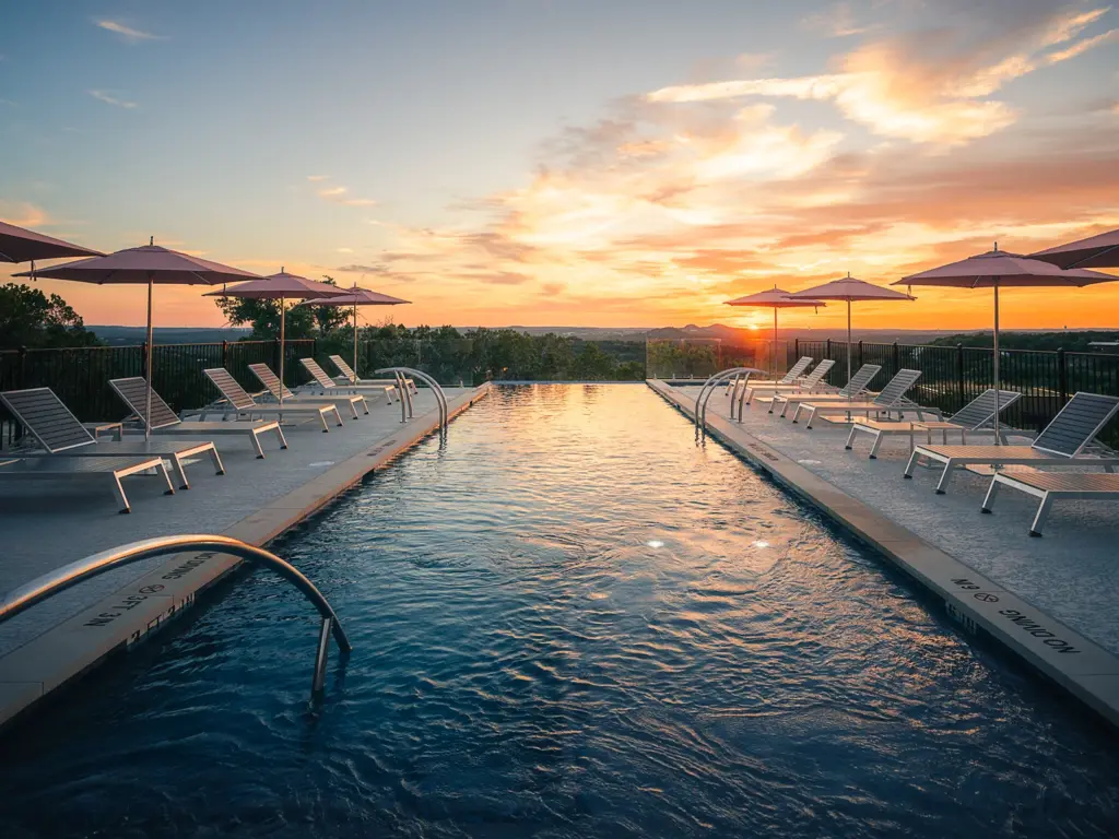 Luxurious infinity pool reflecting a vibrant sunset, lined with modern lounge chairs and pink umbrellas overlooking a vast landscape.