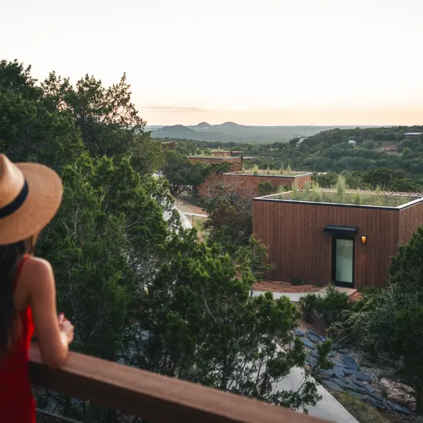 **Onera Wimberly x Paul Clark-7.jpg** > Woman in a red dress and straw hat looking out from a wooden balcony at modern, cube-shaped wooden cabins with green living roofs nestled in a hilly, forested landscape at sunset.
