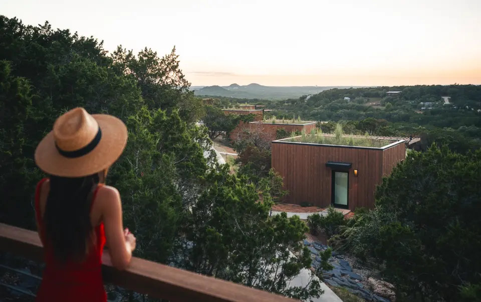 **Onera Wimberly x Paul Clark-7.jpg** > Woman in a red dress and straw hat looking out from a wooden balcony at modern, cube-shaped wooden cabins with green living roofs nestled in a hilly, forested landscape at sunset.