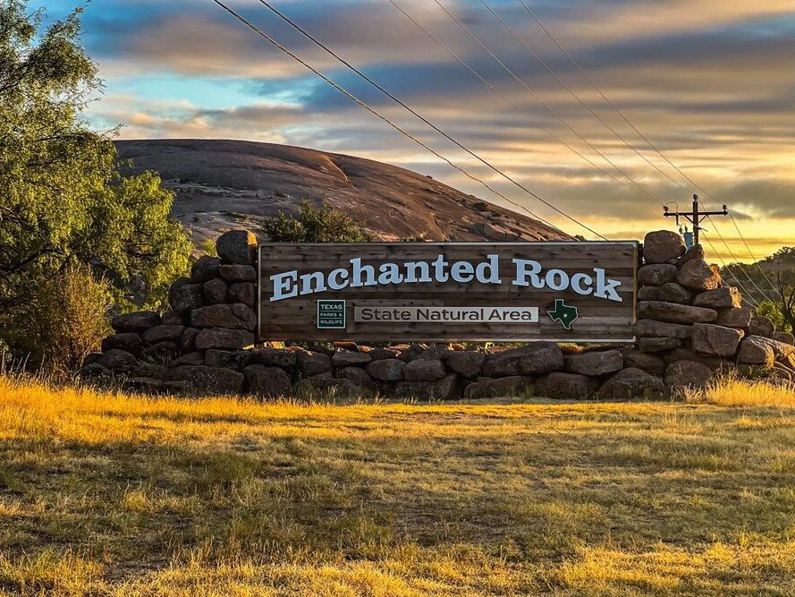 Entrance sign for Enchanted Rock State Natural Area with the large pink granite dome in the background at sunset.