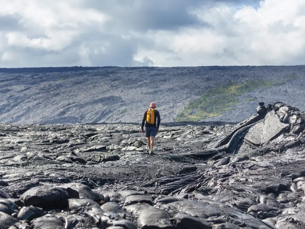 A person walking on black volcanic rock with a yellow backpack and hat, surrounded by a vast landscape.