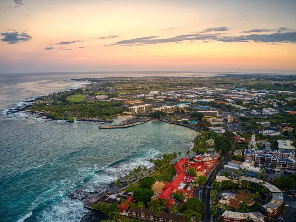 Aerial view of a Kona, with lush greenery, buildings, and ocean waves at sunset. Best time to visit big island