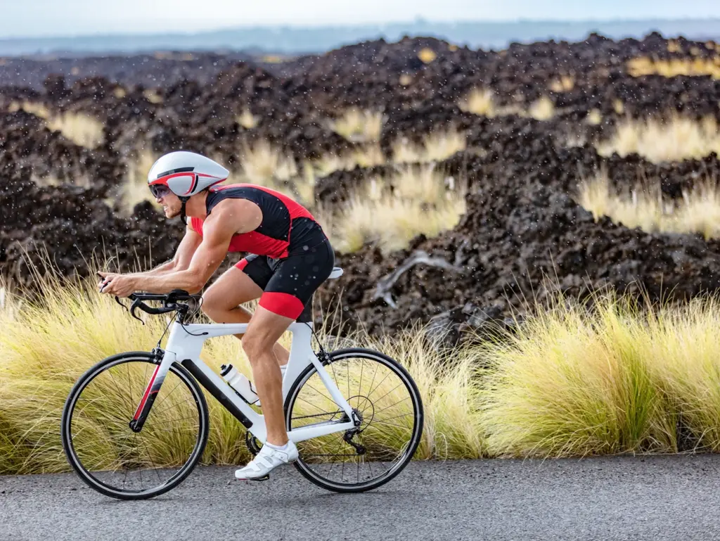 A cyclist in a red and black outfit rides a white bike on a rocky road, near tall grasses.