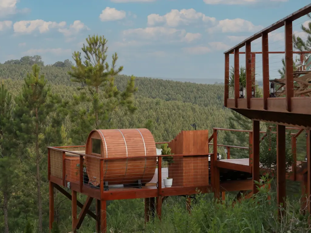 A wooden deck with a barrel sauna and privacy screen overlooking a dense green pine forest landscape.