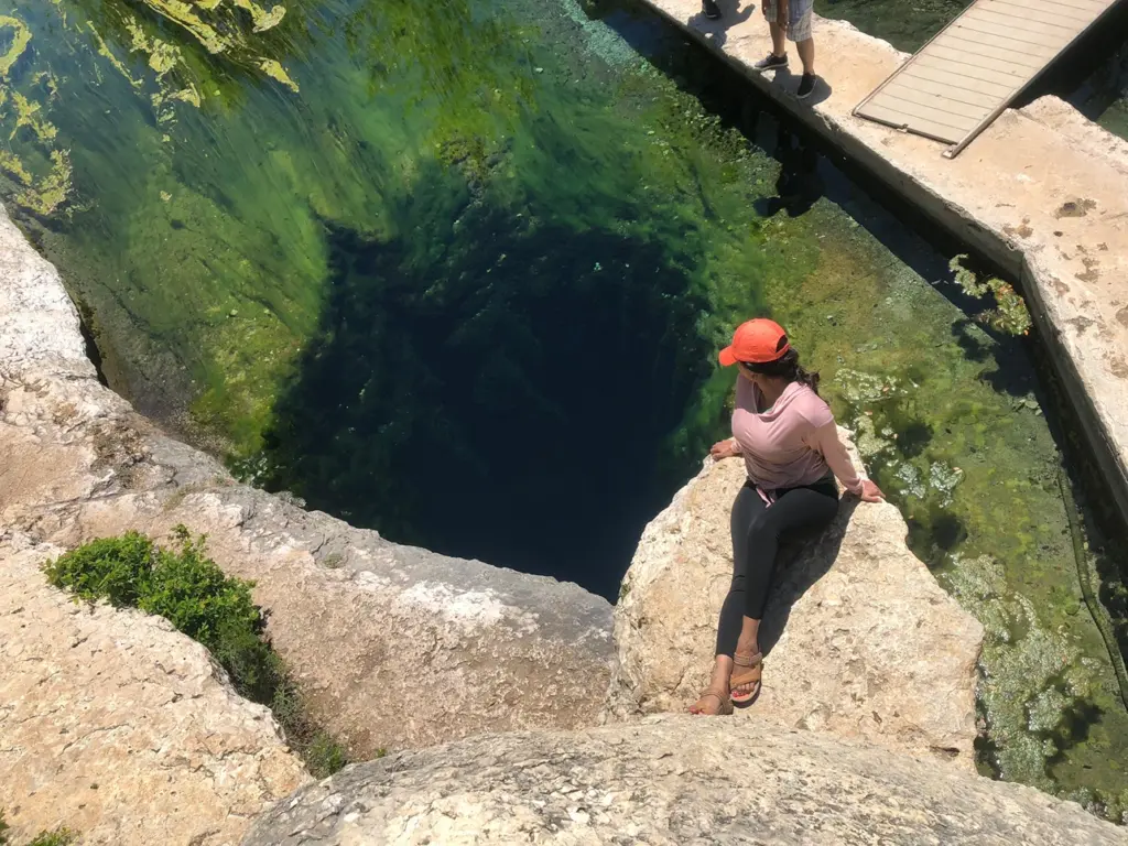 A person sits on limestone rocks peering down into the deep, clear artesian spring at Jacob's Well.