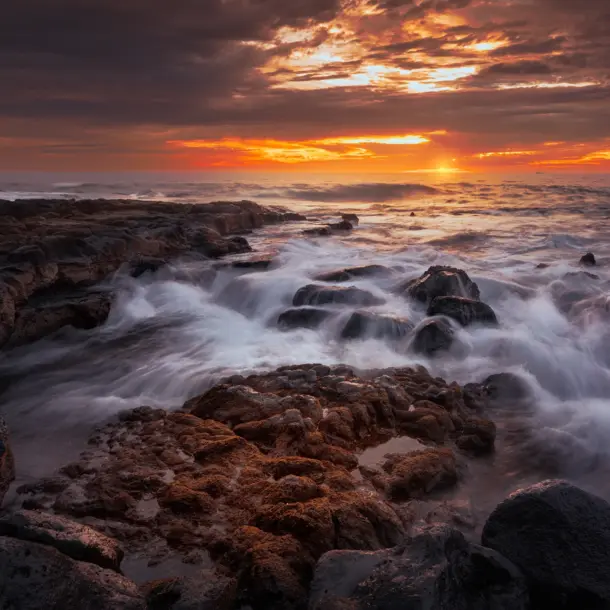Dramatic golden sunset over a rugged volcanic coastline with ocean waves crashing against dark lava rocks.