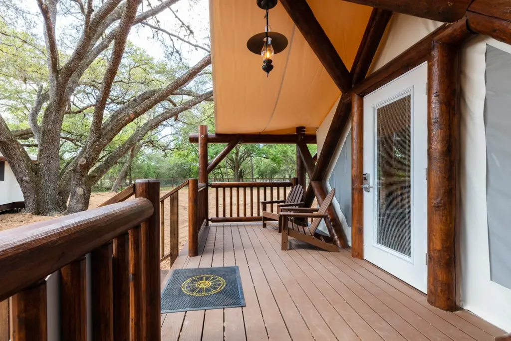 A wooden porch on a canvas tent featuring log railings and two brown Adirondack chairs under a shaded canopy.