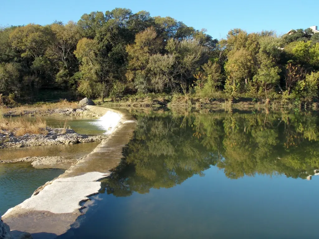 Calm river reflecting dense green trees on the bank next to a low stone dam structure.