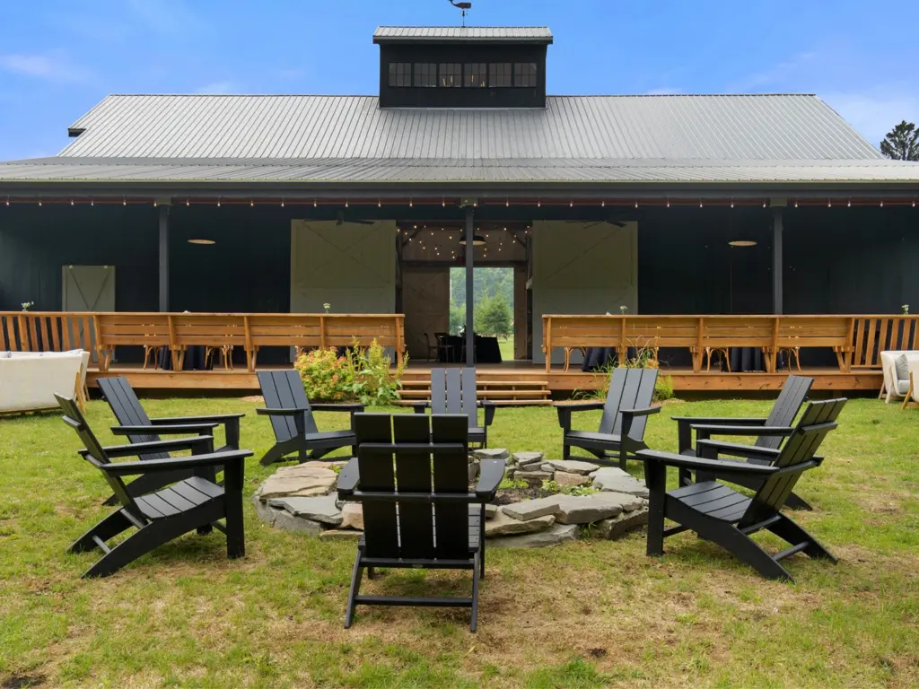 Black Adirondack chairs arranged in a circle around a stone fire pit on a lawn behind a large dark barn.