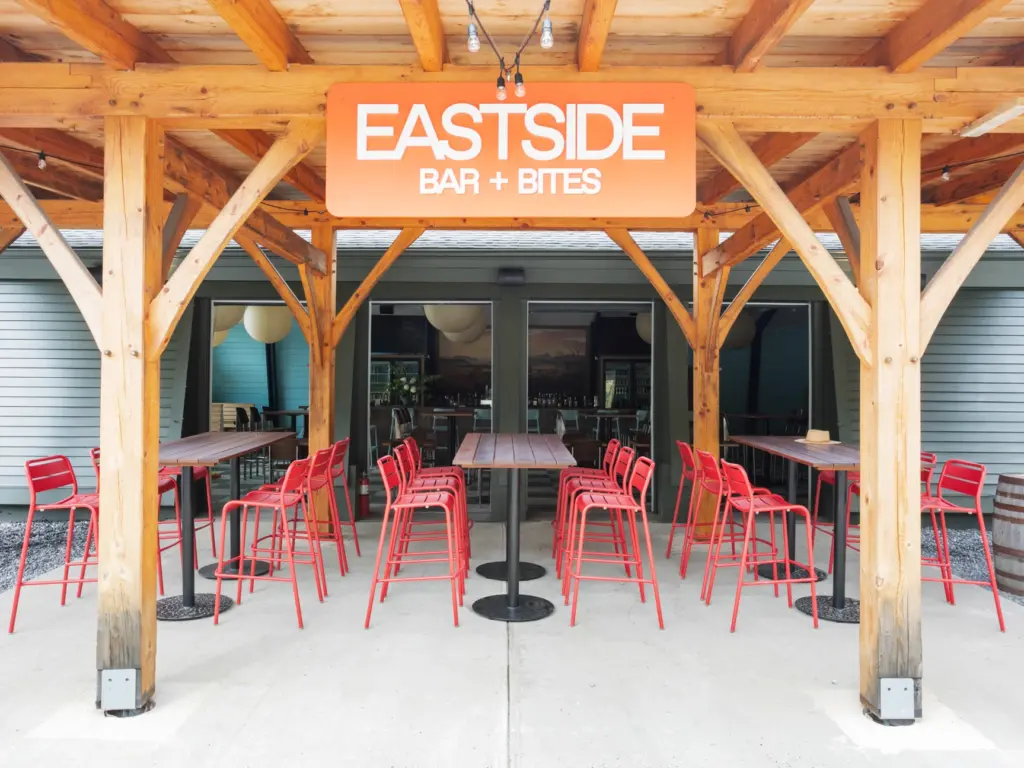 Bright red barstools lined up at an outdoor wooden counter under a pavilion labeled Eastside Bar and Bites.