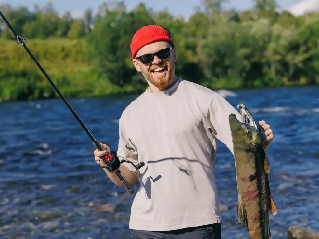 A smiling man in a red beanie holds up a large caught salmon while standing by a flowing river.