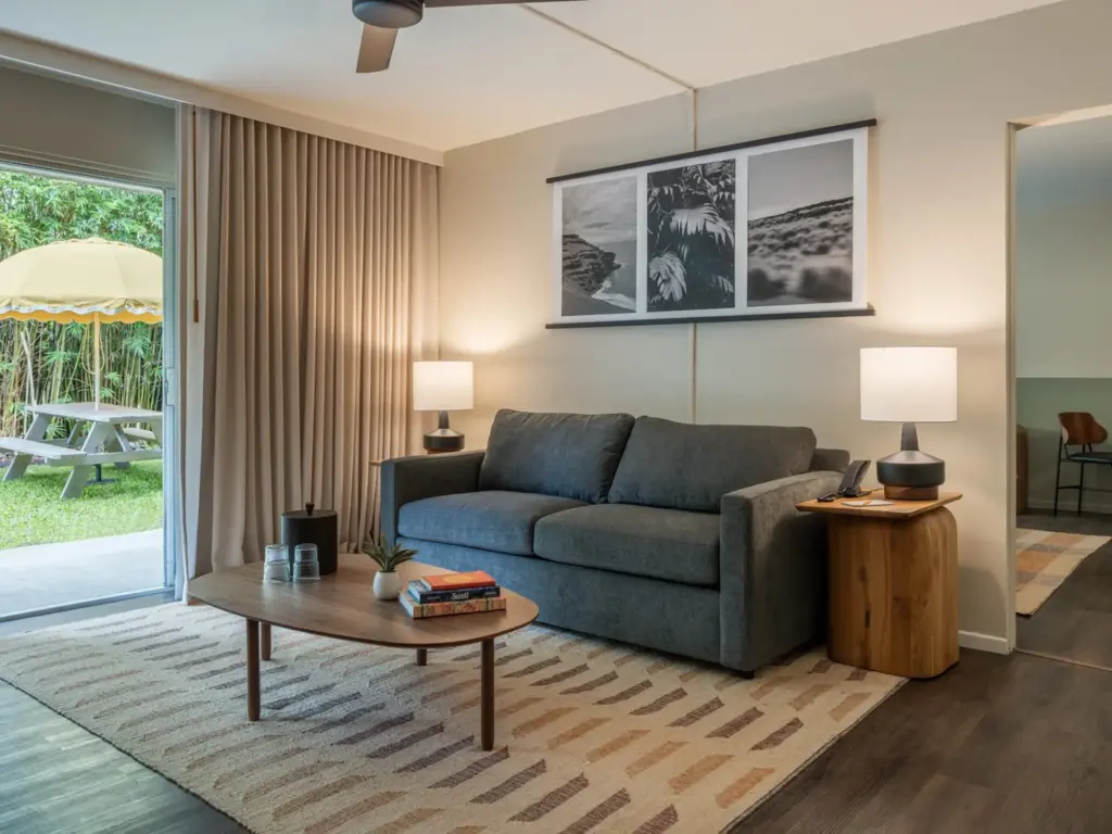 Modern hotel room interior featuring a grey sofa, wooden coffee table, and glass doors to a patio.