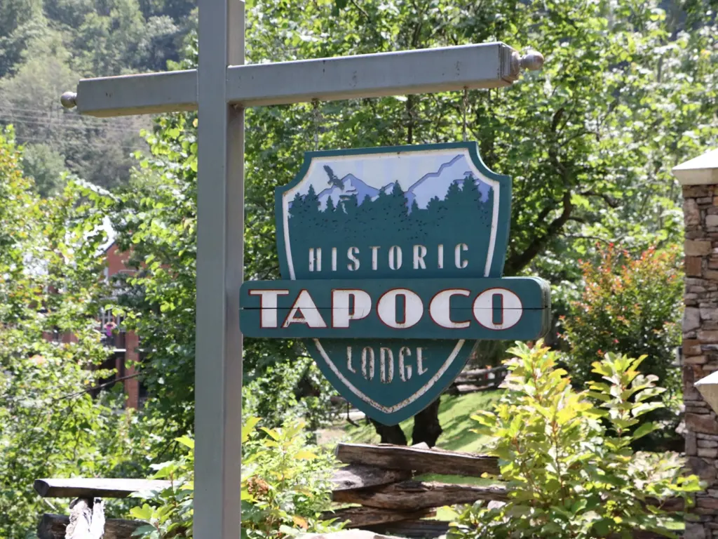 A hanging sign for Historic Tapoco Lodge displayed outdoors against a backdrop of lush green trees.