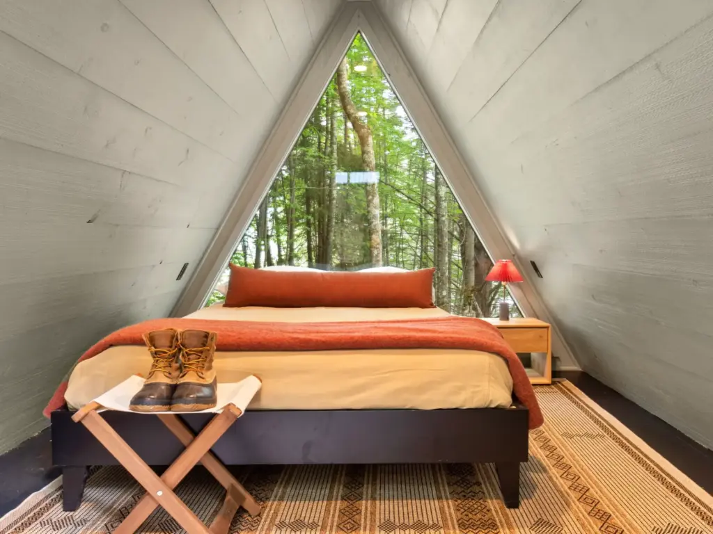 A-frame cabin bedroom with a large triangular window overlooking tall trees, a bed with orange bedding, and hiking boots on a folding stool.