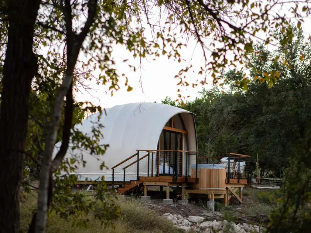 A white arch-roofed glamping tent with a hot tub on a deck among trees, evoking design-forward nature retreats.