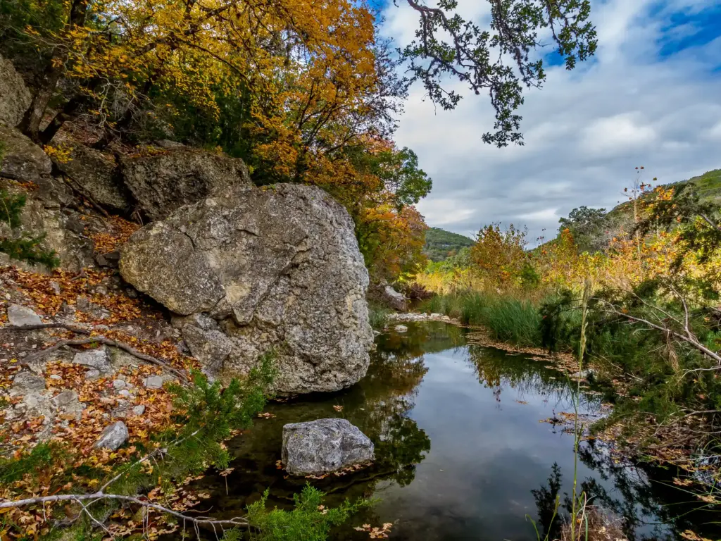 A still creek reflects autumn foliage and cloudy sky beside large granite boulders in a wooded canyon.