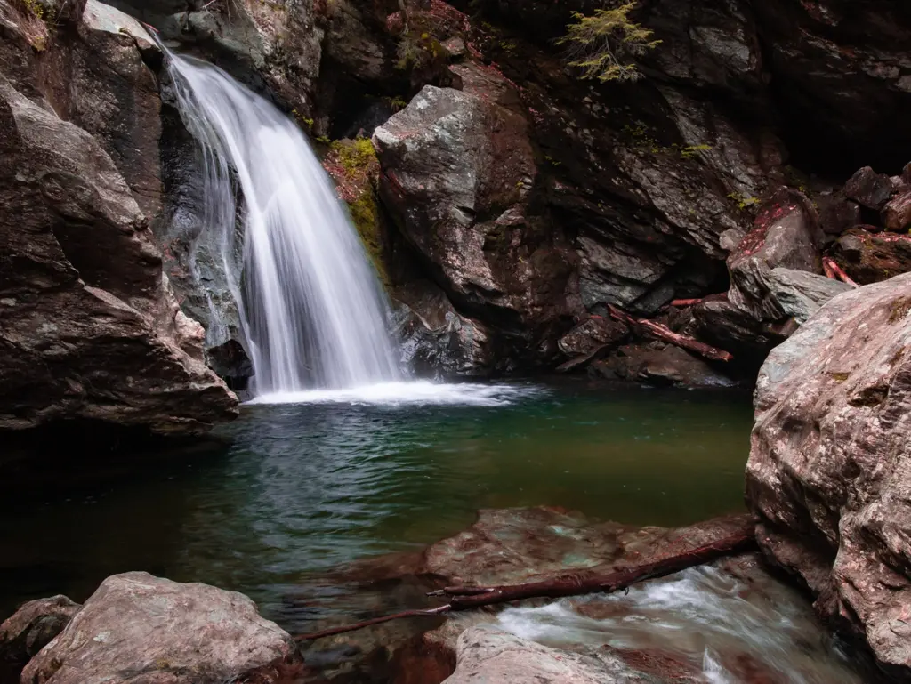 White waterfall tumbling down a steep rocky gorge into a dark green pool at Bingham Falls, Vermont.