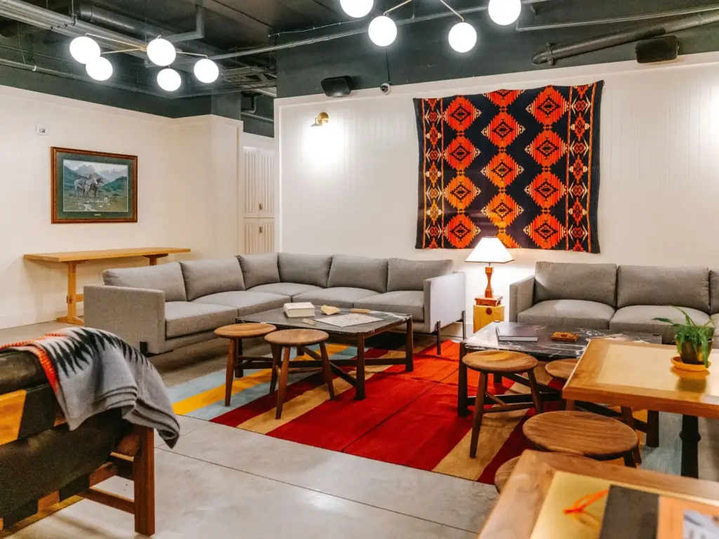 Gray L-shaped sectional sofa surrounds wooden stools and a red rug beneath a bold geometric woven tapestry on a white wall.
