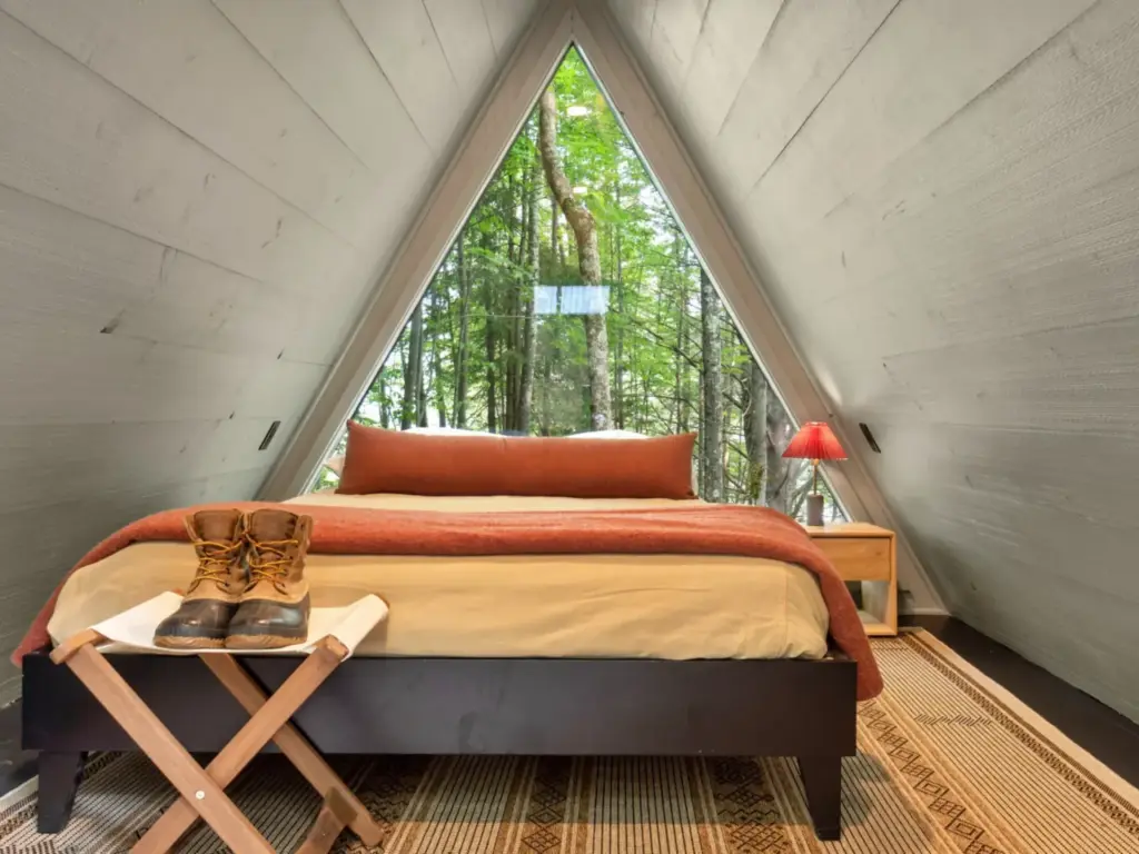 A-frame cabin bedroom with a triangular window overlooking forest trees, a bed with rust blanket, and hiking boots on a stool.