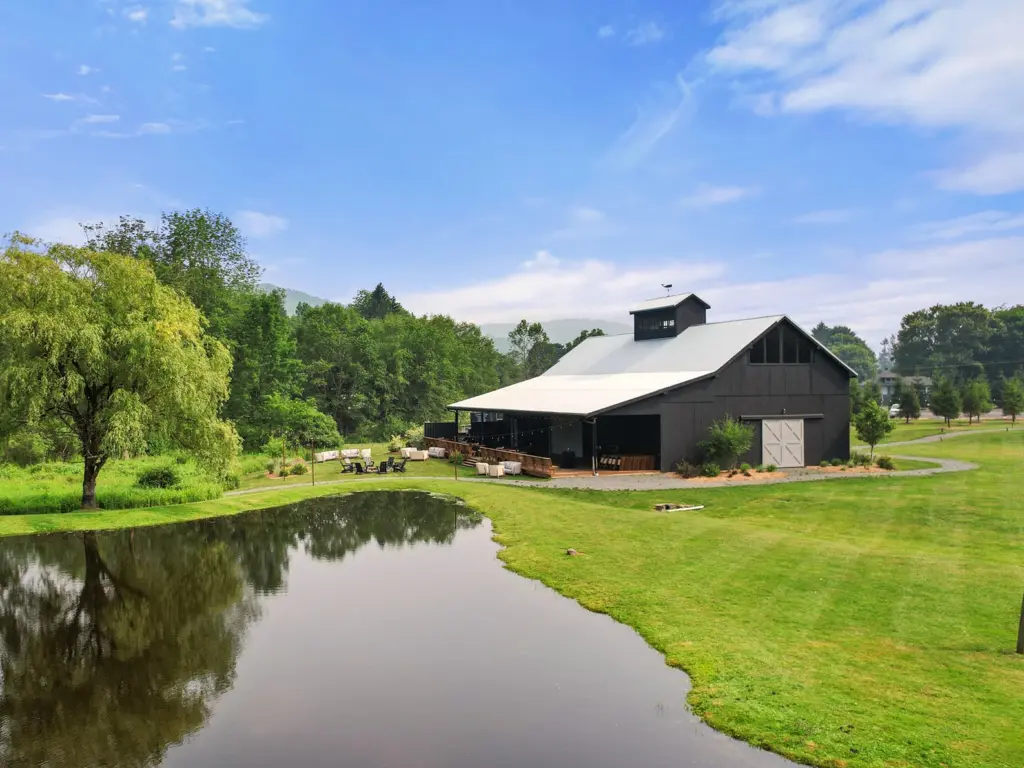 Large black barn next to a calm pond and green lawn, a beautiful destination when choosing where to travel in august.