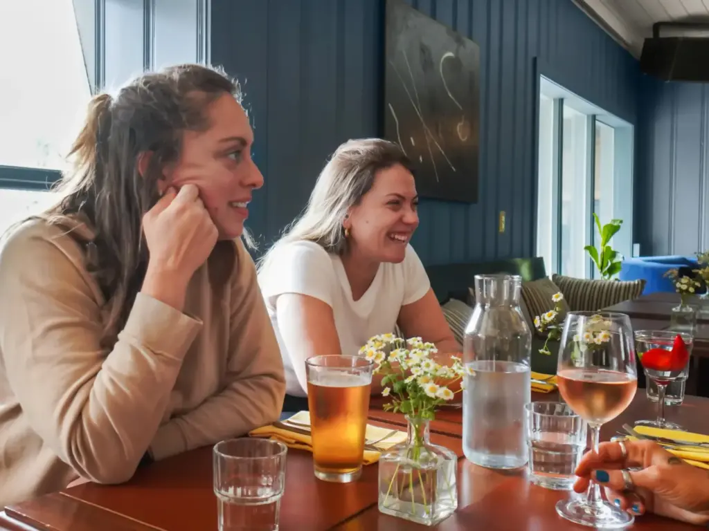Two women laughing together at a restaurant table with drinks and small wildflower vases.