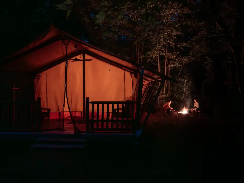 Two people enjoy a campfire beside a canvas tent on a deck, evoking design-forward nature retreats in a wooded setting.