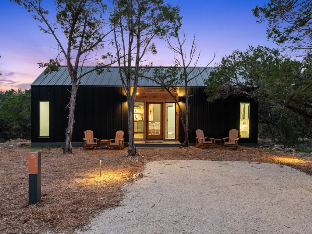 Modern black cabin with glowing windows and wooden patio chairs at dusk at The Cedars Ranch.