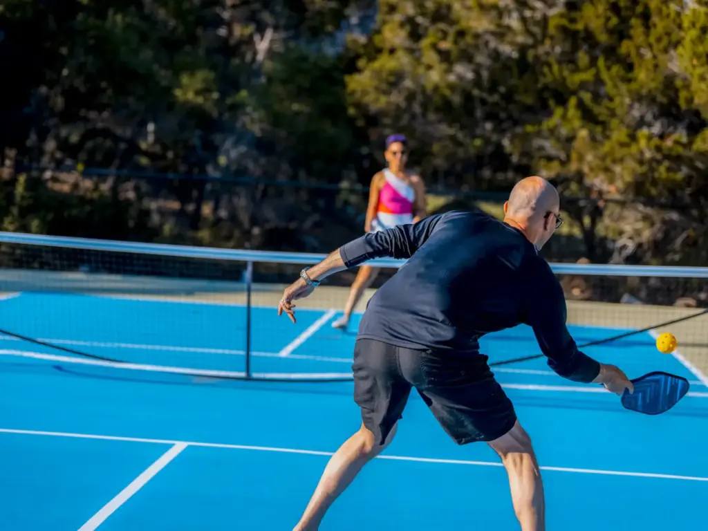 Two people playing pickleball on a blue outdoor court, with a man reaching to hit a yellow ball.
