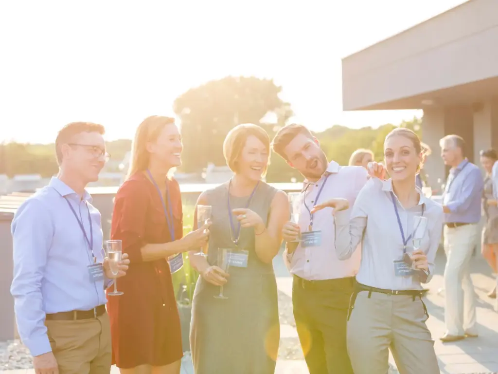 Five professionals with lanyards laughing and holding champagne glasses at an outdoor rooftop networking event at sunset.