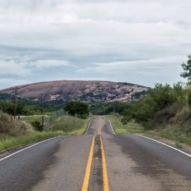 Two-lane highway on one of the best road trips in Texas, leading toward a pink granite dome above Hill Country scrubland.