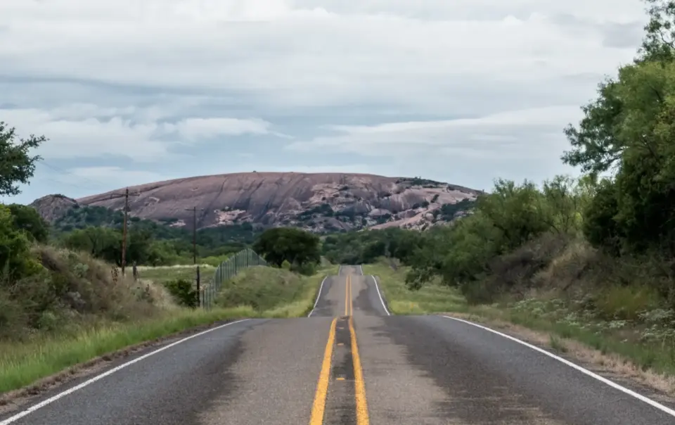 Two-lane highway on one of the best road trips in Texas, leading toward a pink granite dome above Hill Country scrubland.