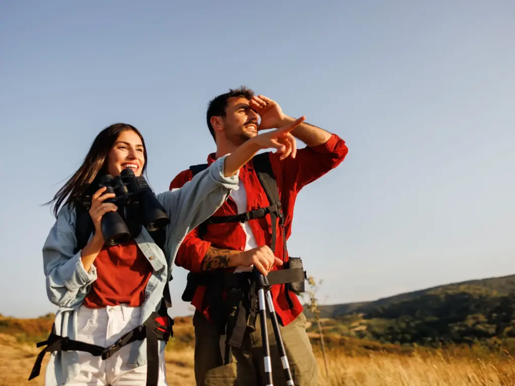 Couple hiking in a sunny field where the woman looks through binoculars and the man points ahead.