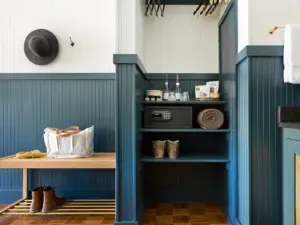 Blue beadboard built-in shelving unit with a digital safe, rolled blanket, glassware, and boots beside a wooden bench with a tote bag.