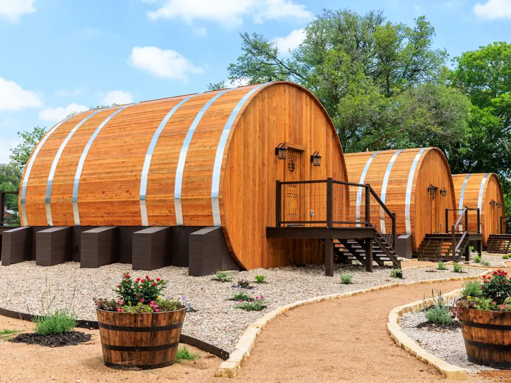 Three large wooden barrel-shaped cabins sit along a gravel walking path surrounded by trees.