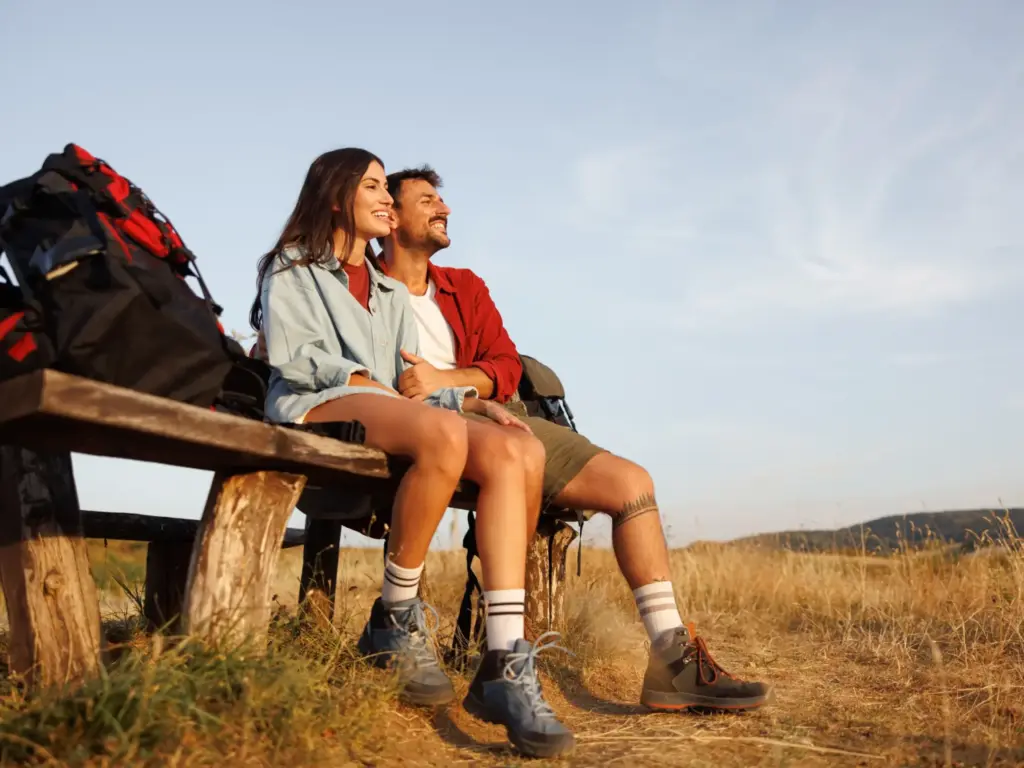 Smiling couple with backpacks resting on a wooden bench, looking for inspiration on where to travel in august.