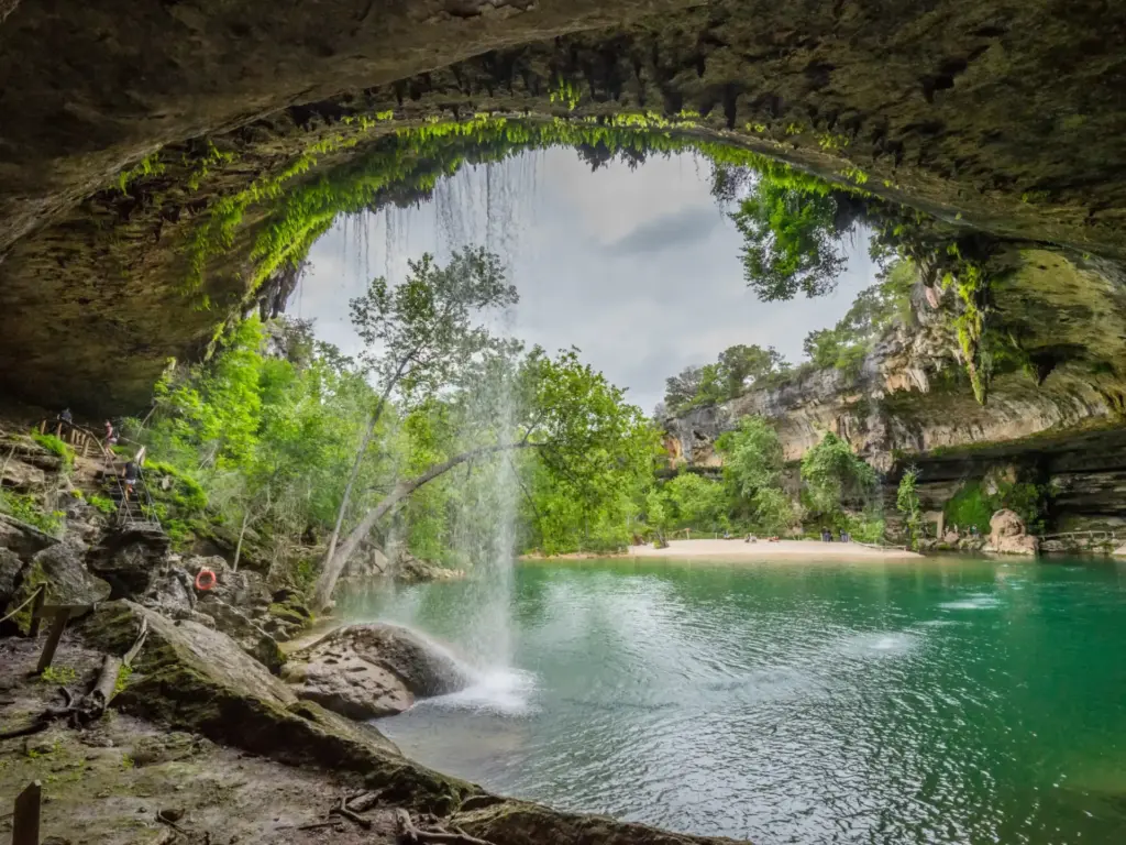 A waterfall cascades into a turquoise swimming hole beneath a limestone arch draped with ferns and trees.