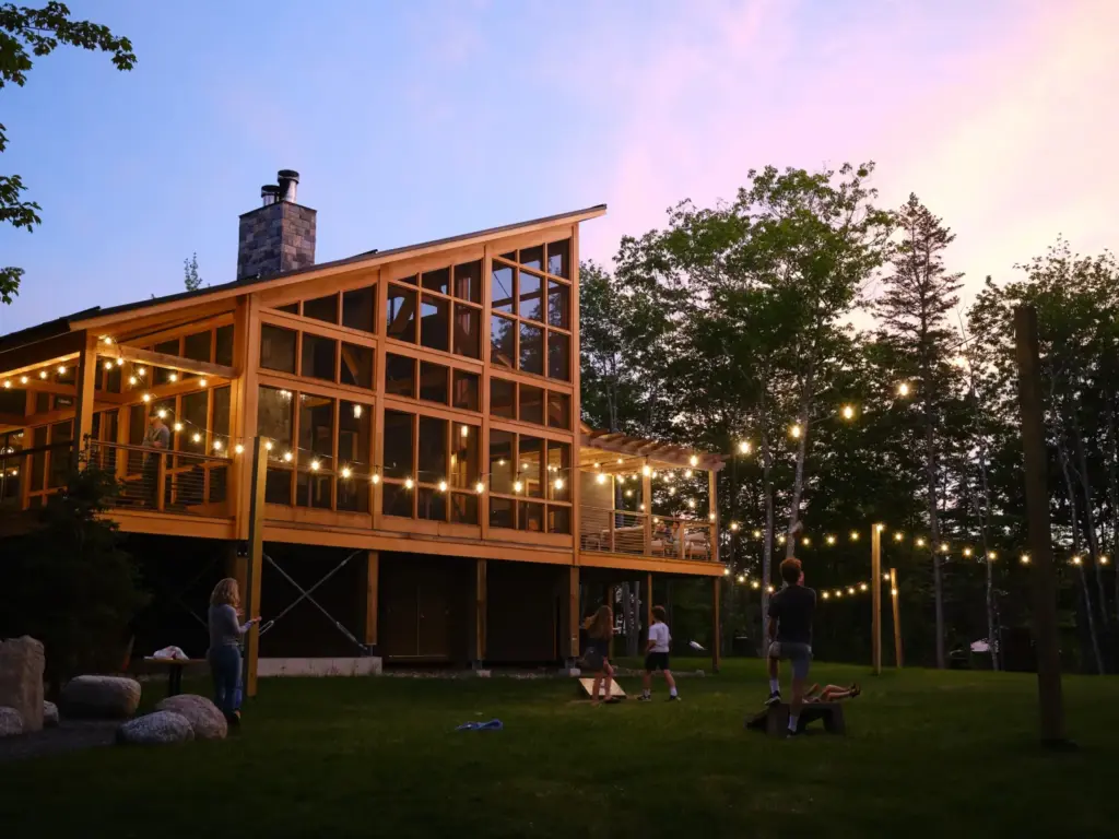 Colleagues gather on a lawn at dusk beneath string lights outside a glass lodge during a nature immersive corporate offsite.
