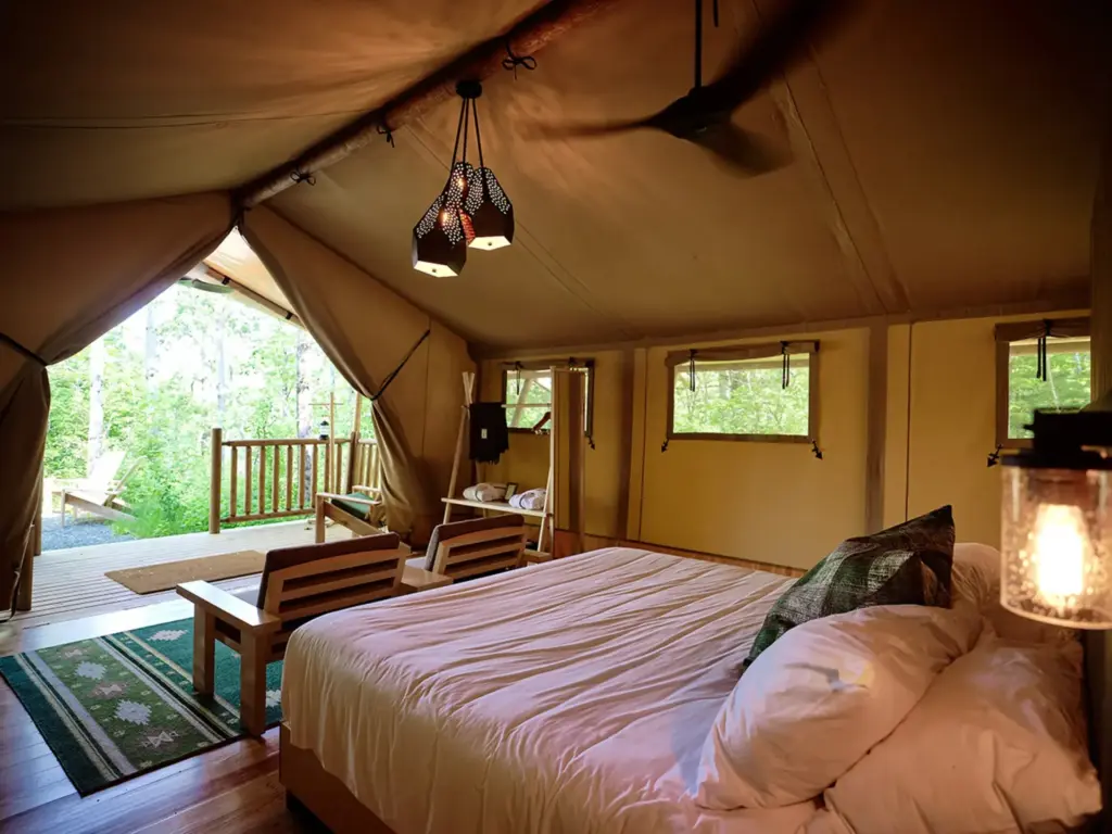 Spacious glamping tent interior with a queen bed, wood floors, hanging lanterns, and an open canvas wall facing a forested deck.