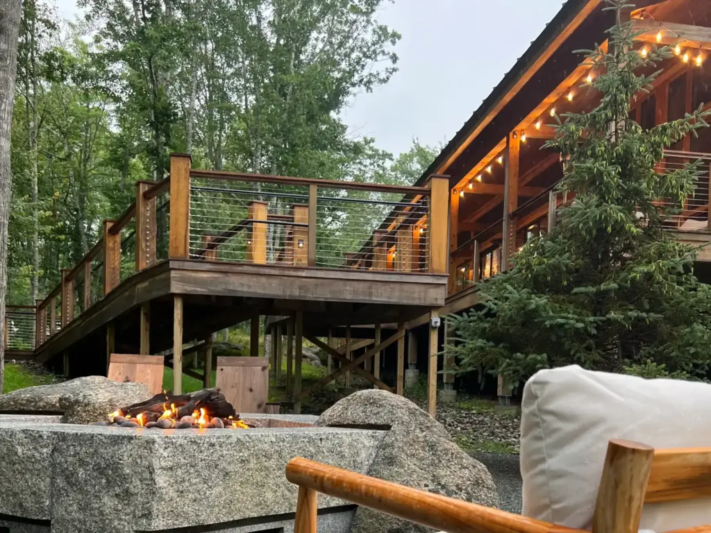 Stone fire pit and cabin deck at one of many design-forward nature retreats, glowing with string lights among trees at dusk.