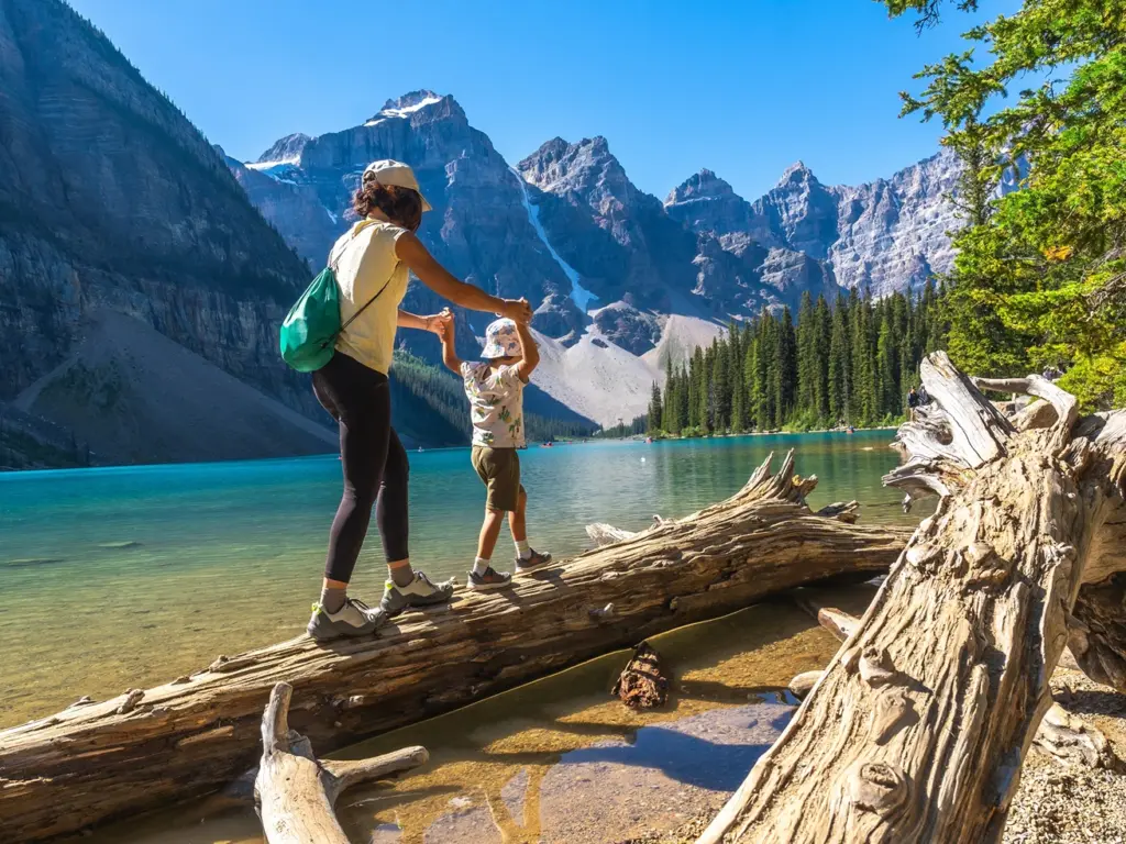 Woman and child walk on a log over a vibrant blue mountain lake, a great choice for where to travel in august.