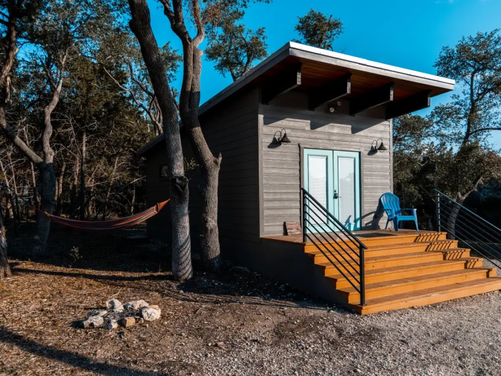 Small modern cabin with gray wood siding, teal double doors, wooden deck stairs, and a hammock strung between trees.