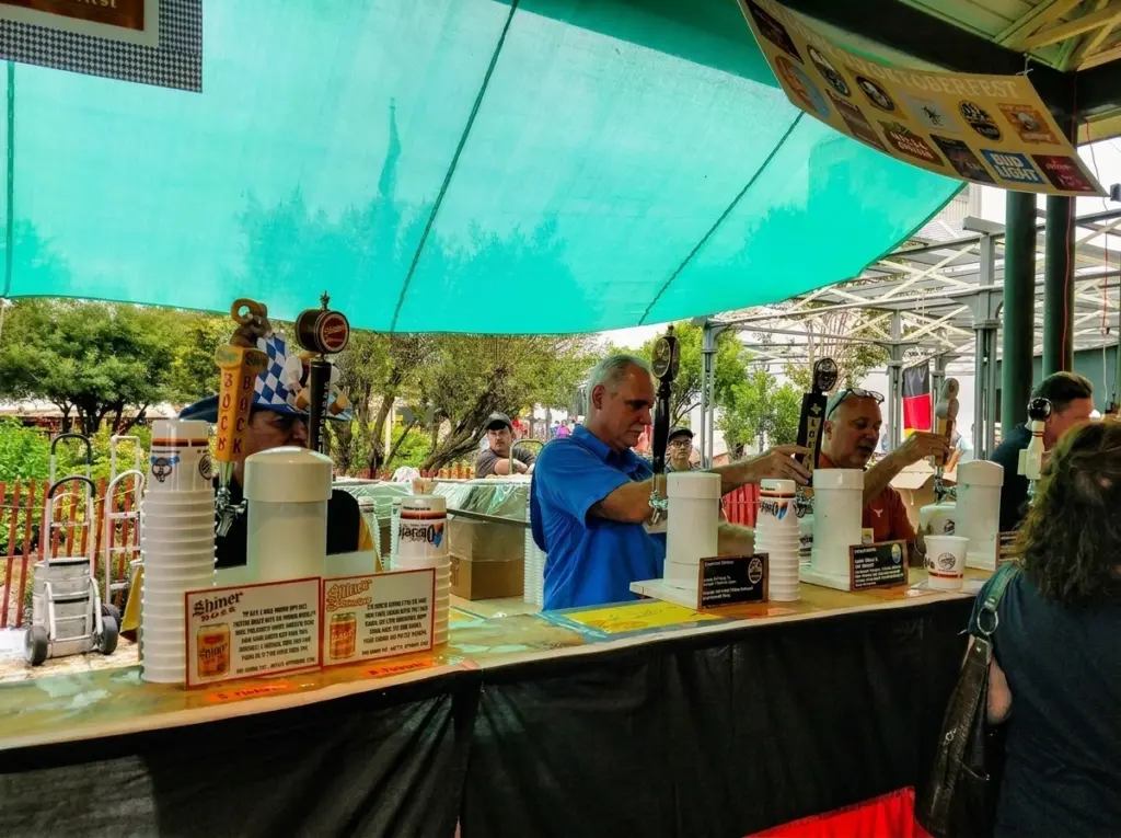 Bartenders serve drinks from taps behind a wooden counter under a green canopy at Fredericksburg Oktoberfest.