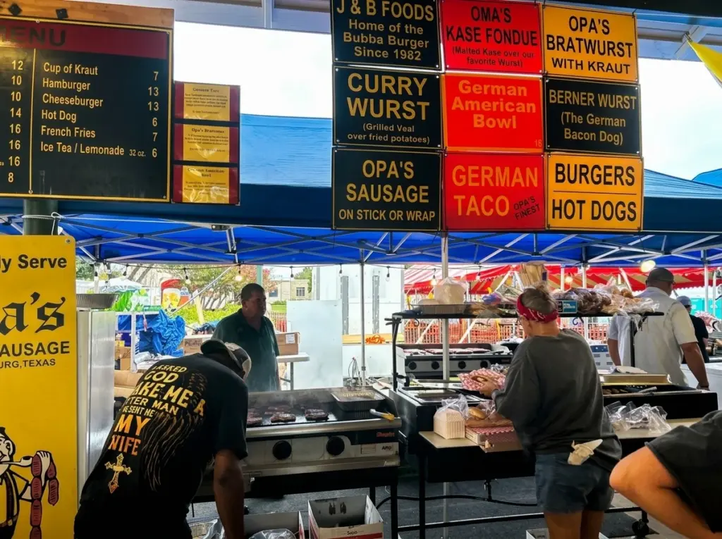 Food vendors cook on a grill under a tent displaying colorful menu signs for bratwurst and other German foods.