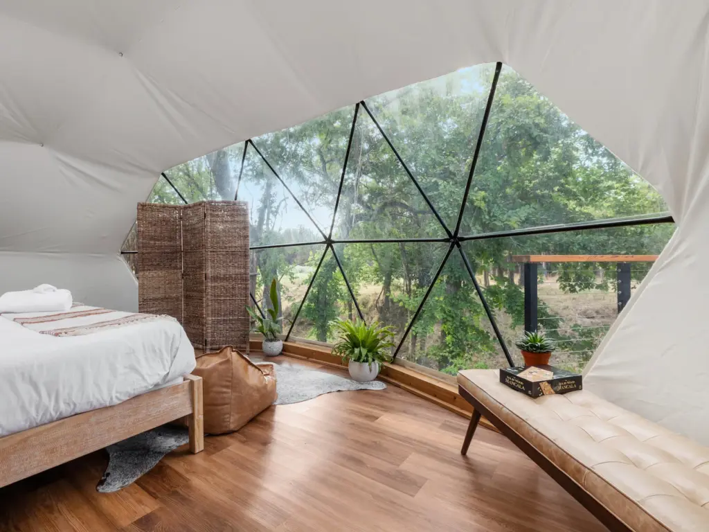 Geodesic dome glamping interior with wood floor, bed, lounge chair, and panoramic triangular window overlooking trees.