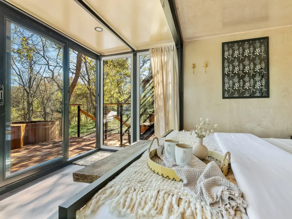A bedroom looking out through glass sliding doors to a forest view and a deck with a soaking tub.