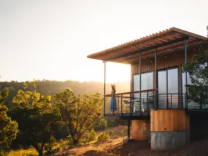 Woman in a blue dress and hat stands on the elevated deck of a modern wood and steel cabin at golden hour, overlooking a wooded hillside. — design-forward nature retreats