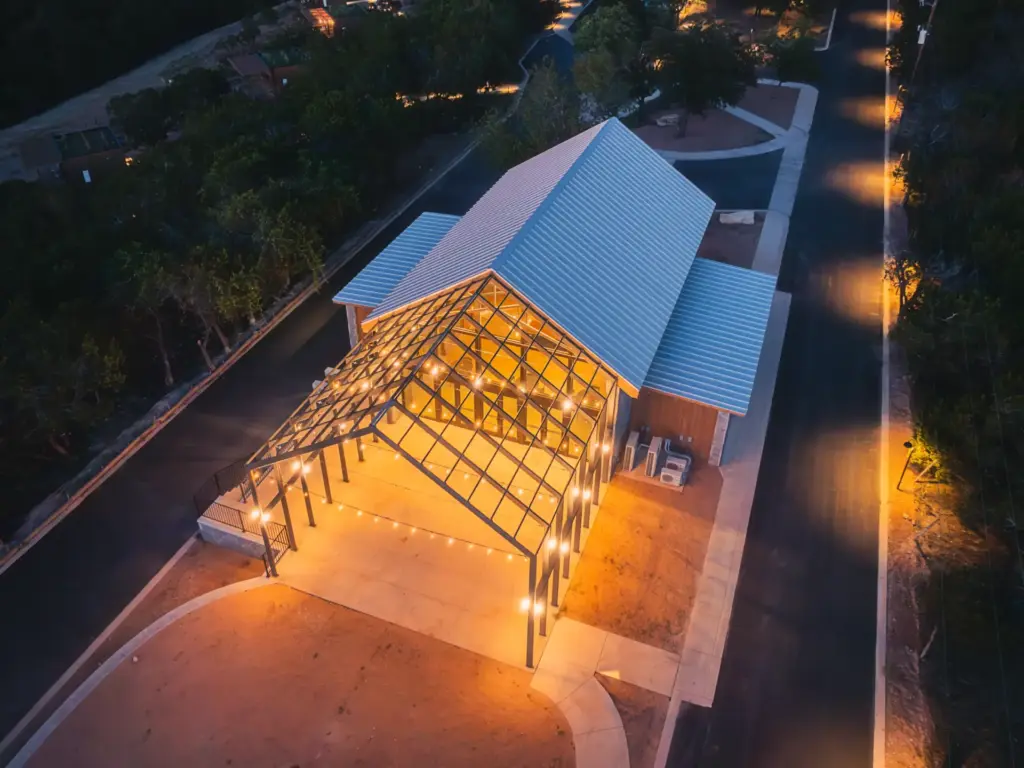 Aerial dusk view of an open-air steel-framed pavilion with string lights beside a building with a metal roof. — design-forward nature retreats