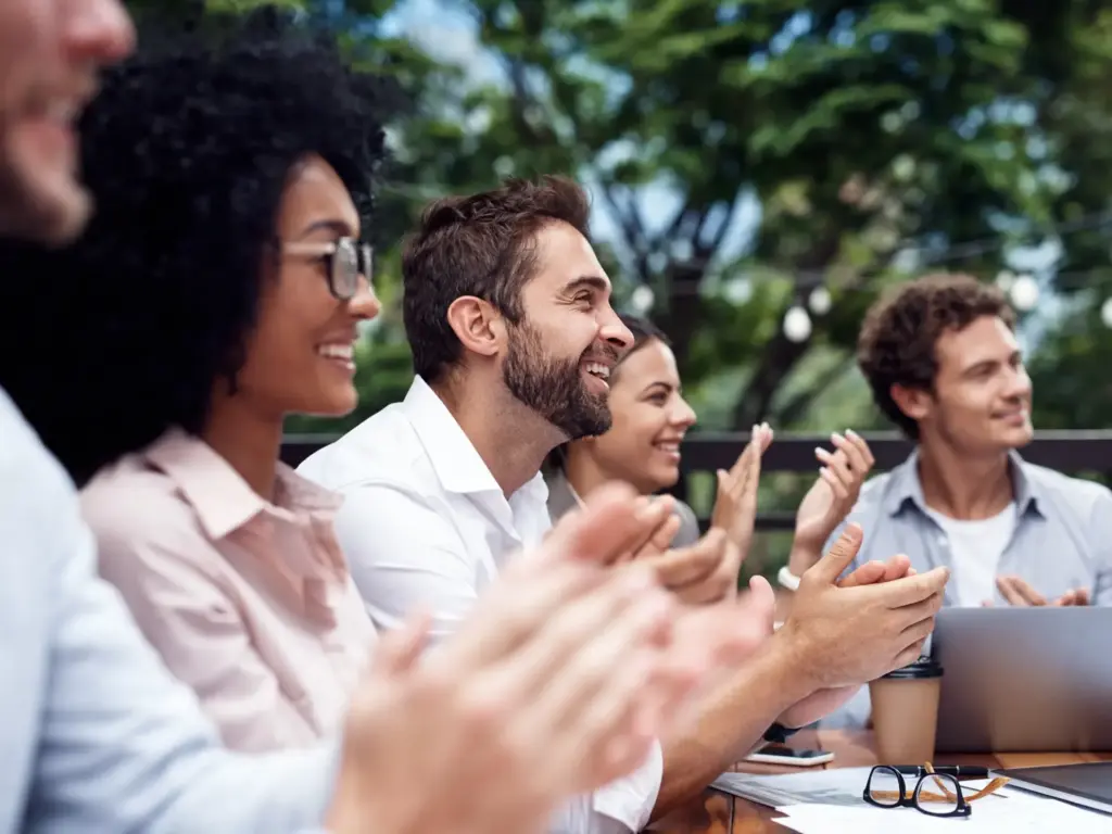 Diverse group of smiling professionals clapping at an outdoor meeting seated at a table.