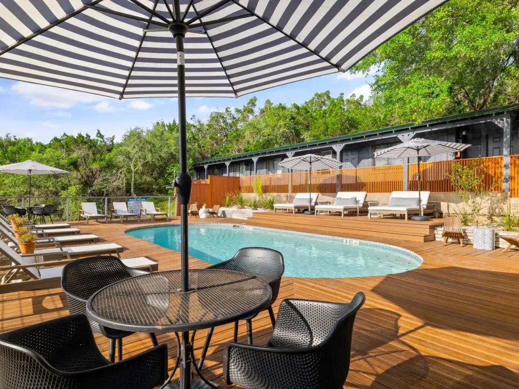 Outdoor pool with wood deck, black mesh patio furniture, striped umbrella, and lounge chairs surrounded by trees.
